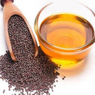 Glass bowl of mustard oil next to wooden scoop with black mustard seeds on white background