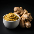 White bowl filled with ginger paste next to fresh whole ginger roots on black background