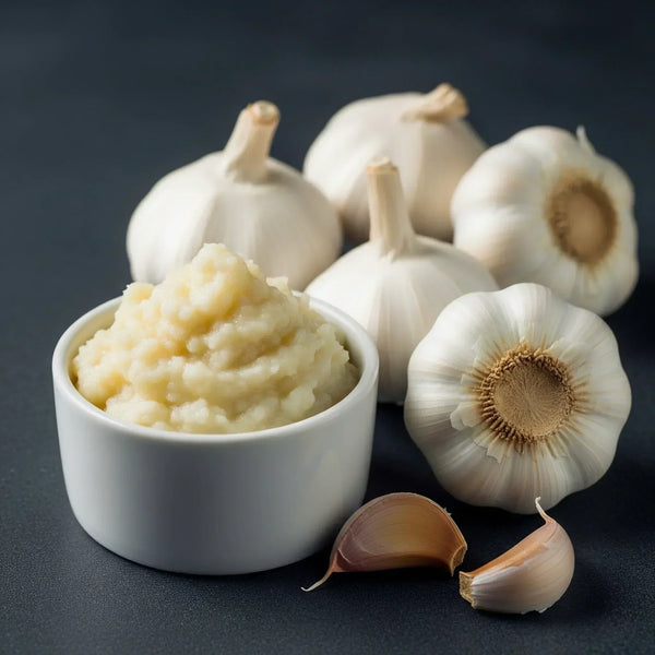 White ceramic bowl with creamy garlic paste surrounded by whole garlic bulbs and cloves on dark surface