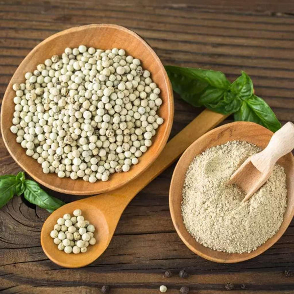 Wooden bowls and spoon with white peppercorns and white pepper powder on wooden surface with basil leaves
