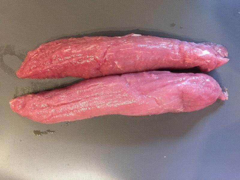 Two raw pink pork tenderloin pieces on a dark cutting board ready for cooking