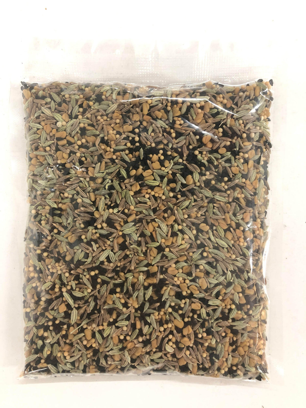 Clear plastic bag filled with mixed whole spices including mustard seeds, fennel seeds, and cumin seeds on white background
