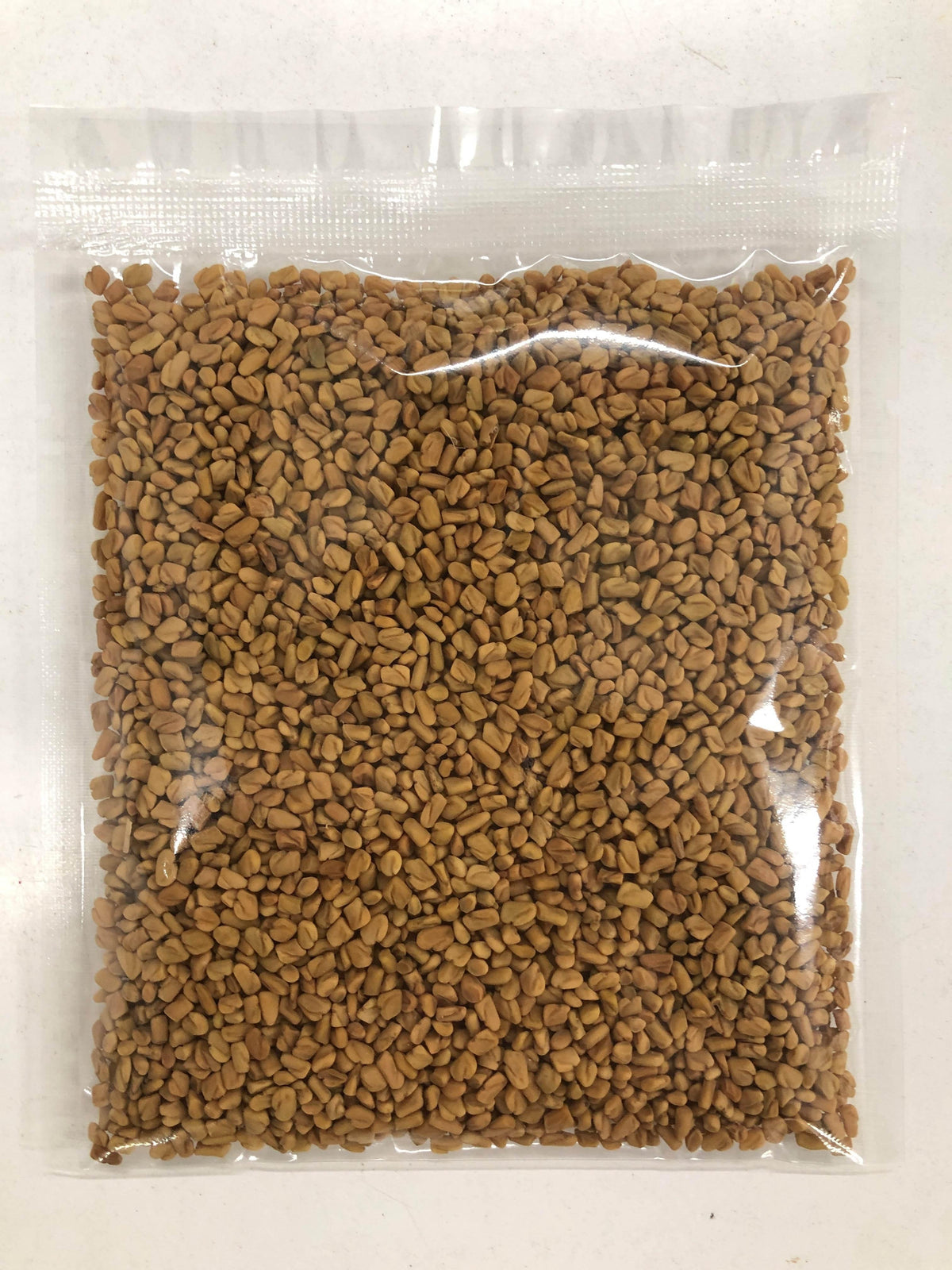 Clear plastic bag filled with small brown fenugreek seeds on a white surface